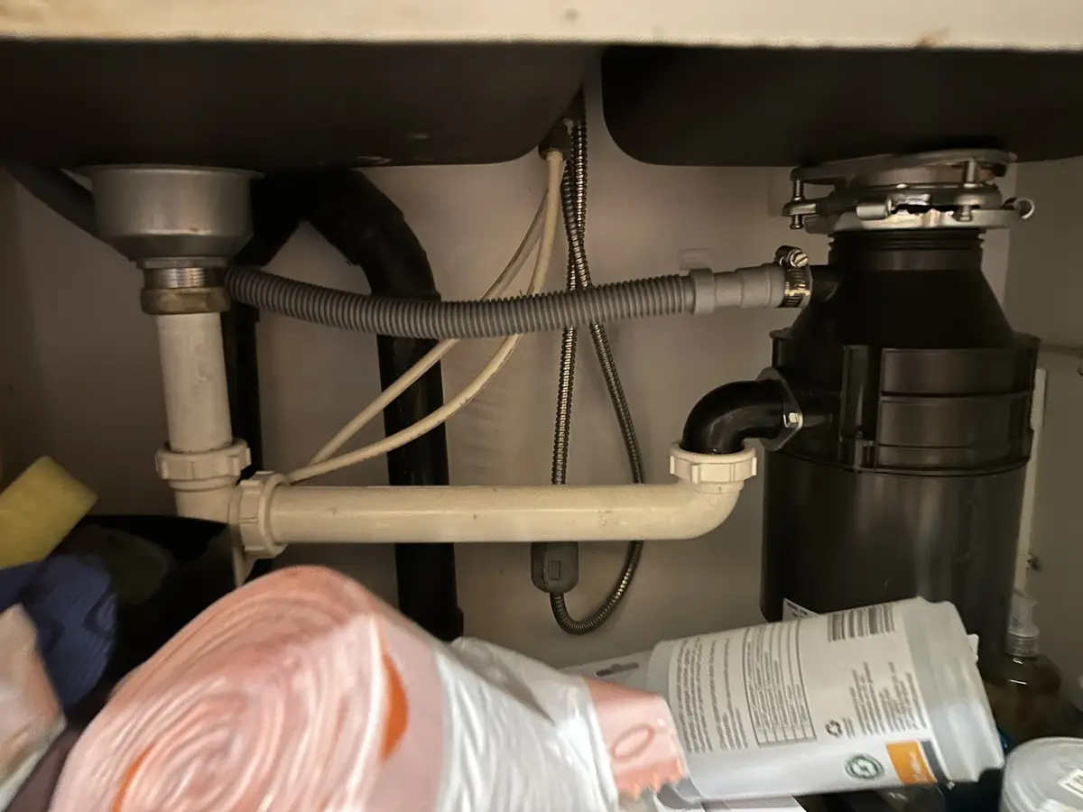 A close-up photo under a kitchen sink showing plumbing, a garbage disposal unit, and various cleaning supplies. The overlaid…