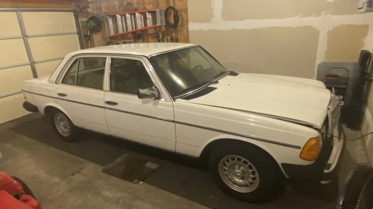A white 1979 Mercedes-Benz 300D diesel sedan is parked inside a garage with unfinished drywall walls and tools hanging on…