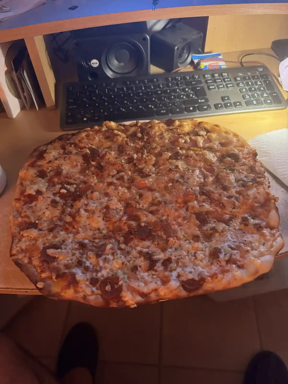 A large, cheesy pepperoni pizza sits on a desk, obscuring a keyboard and speakers behind it. The pizza is so big it extends…