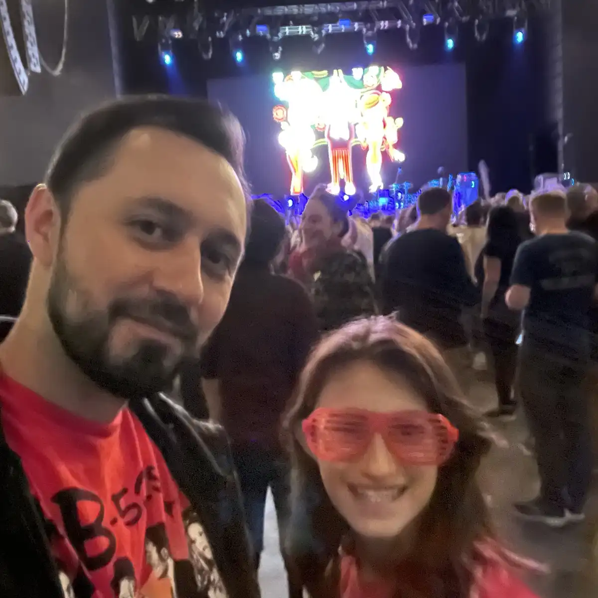 A selfie of a man and a woman at a concert, with the woman wearing bright pink novelty sunglasses and both smiling. Behind…