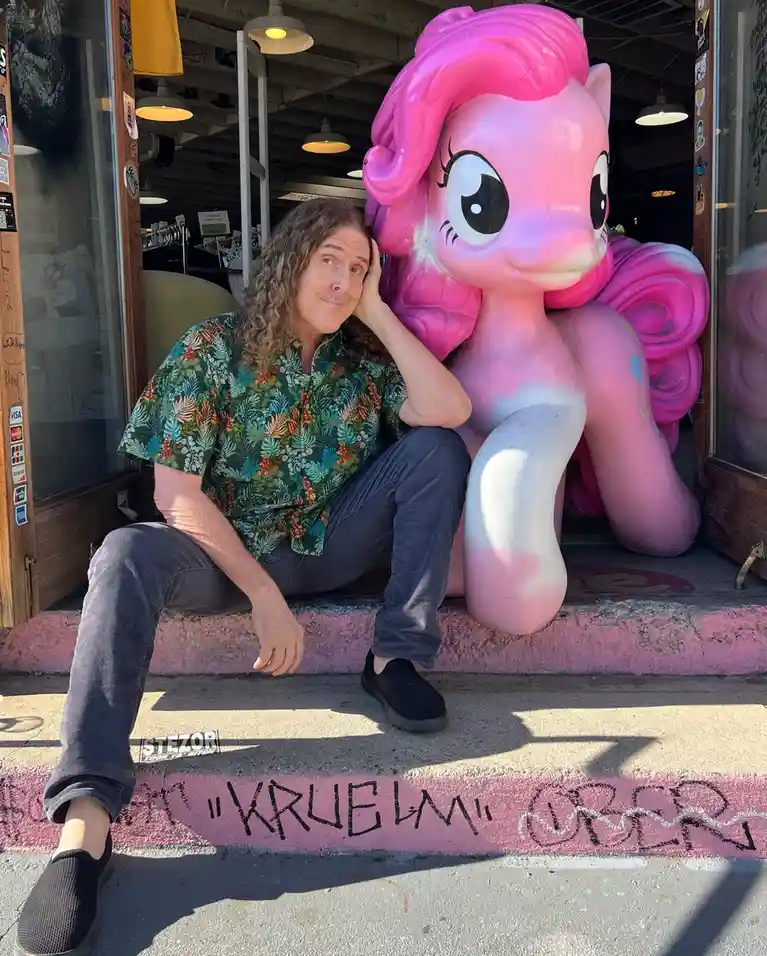 A man with long curly hair sits on a pink-painted step next to a large, pink, inflatable statue of the cartoon character…