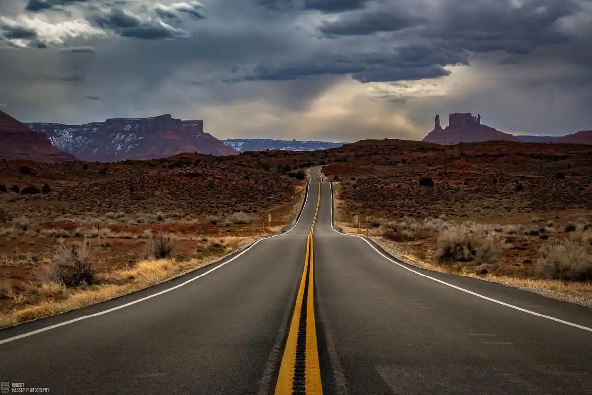 A dramatic desert landscape features a long, straight asphalt road stretching toward distant red rock formations under a…