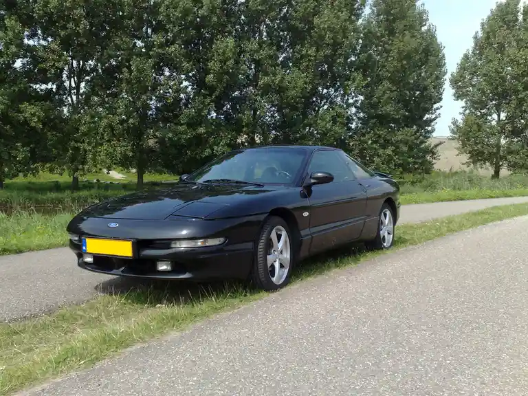 A black Ford Mustang convertible is parked on the grass beside a paved road, surrounded by green trees and grass under a…