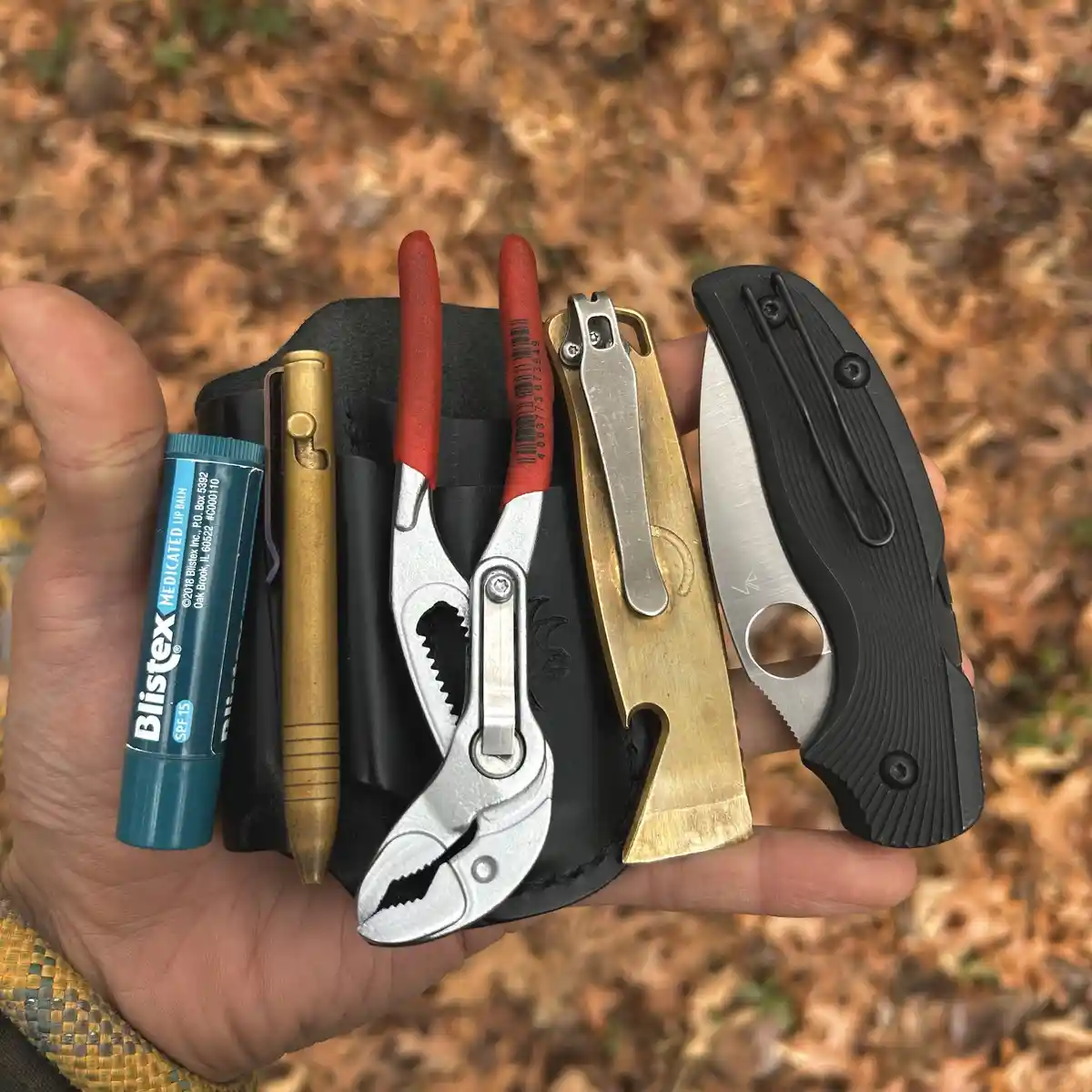 A hand holds a black leather sheath containing an assortment of tools including a Blistex lip balm, a brass pen, red-handled…