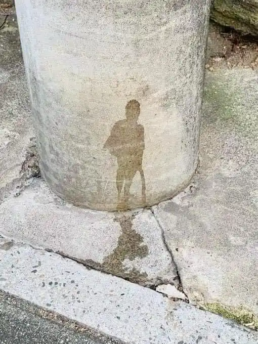 A weathered concrete pillar shows a faint, brownish stain resembling a human figure standing upright. The stain is located…