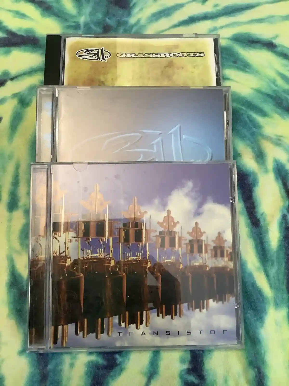 Three 311 CD cases are stacked vertically on a tie-dye fabric background. The bottom case shows the album 'Transistor' with…
