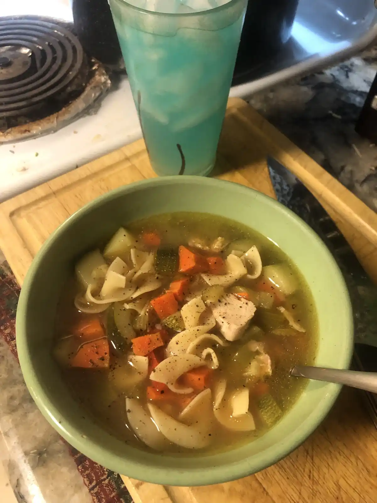 A close-up photo shows a light green bowl of homemade chicken noodle soup with visible noodles, carrots, chicken pieces, and…