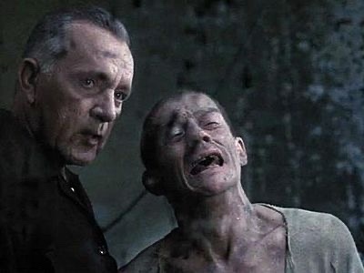 A still from a movie shows an older man in a dark jacket looking intently at a younger, emaciated man with a distressed…
