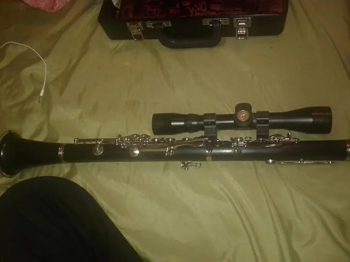 A clarinet lies horizontally on a green bedspread, with a black rifle scope mounted on top of it. In the background, a black…