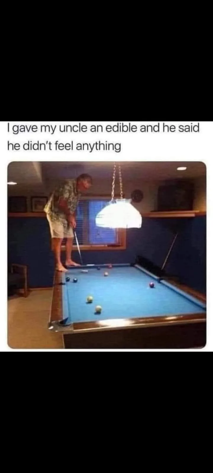 A man in a patterned shirt and shorts stands barefoot on the edge of a pool table, leaning over to take a shot with a cue…