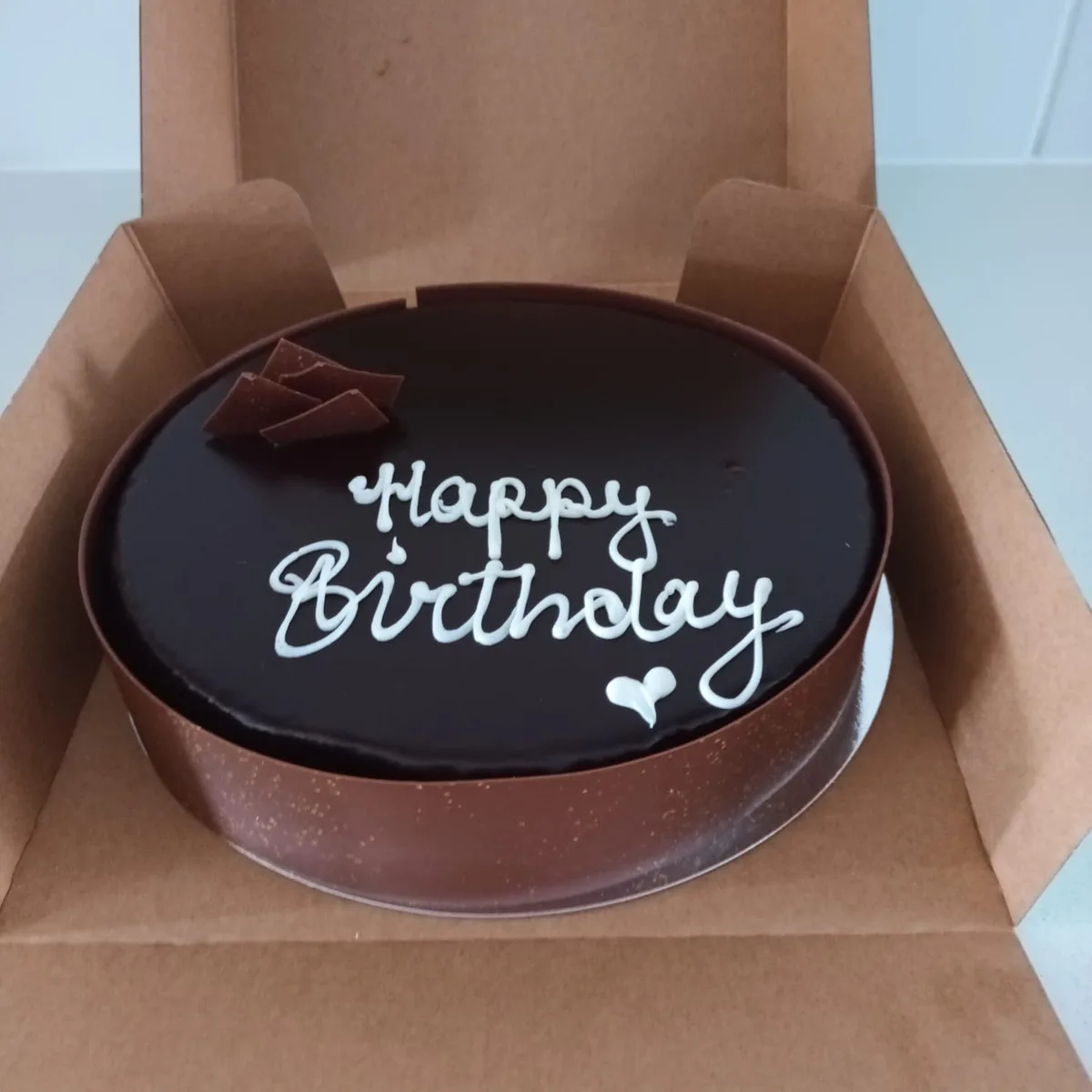 A round chocolate cake with smooth dark chocolate frosting sits inside an open brown cardboard box. The cake is decorated…