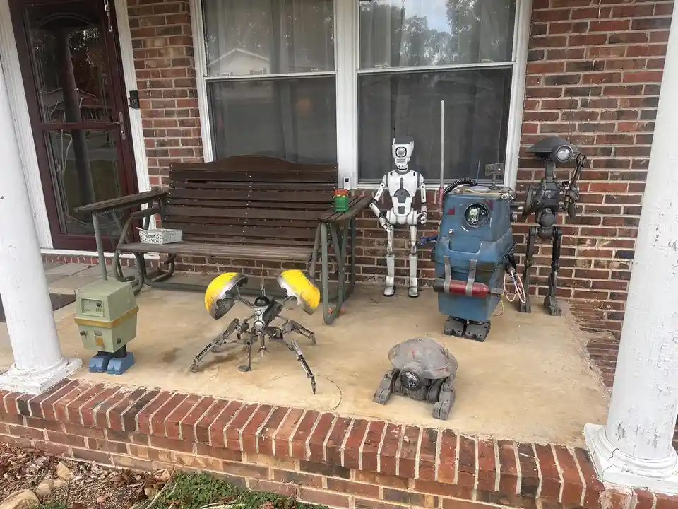 A front porch with a wooden bench and brick walls features several homemade robot figures, including a white humanoid, a…