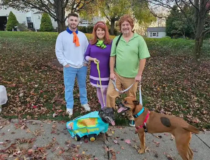 Three people dressed as Scooby-Doo characters (Fred, Daphne, and Shaggy) stand outdoors with two dogs. One dog wears a…