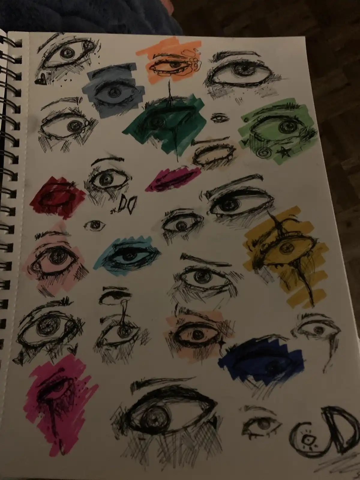A sketchbook page filled with numerous hand-drawn eyes in various styles, some colored with markers in red, blue, green…