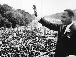 A black and white historical photograph shows Dr. Martin Luther King Jr. waving to a massive crowd from a raised platform…
