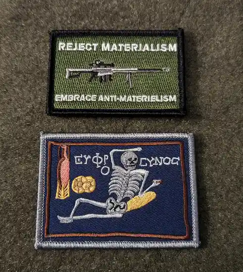 Two embroidered patches lie on a gray fabric surface. The top patch is olive green with white text reading 'REJECT…