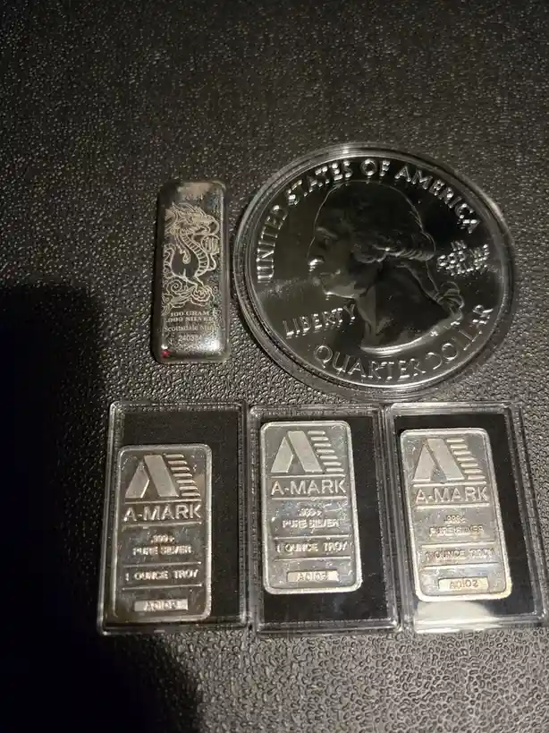 A photo shows several silver items on a textured black surface: a small dragon-themed silver bar, a U.S. quarter dollar coin…