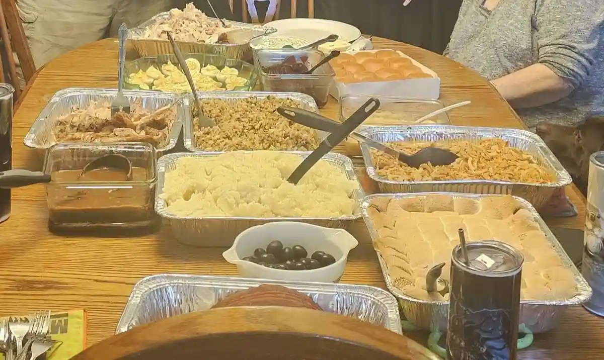 A wooden dining table is covered with numerous aluminum trays and glass dishes filled with various holiday foods including…