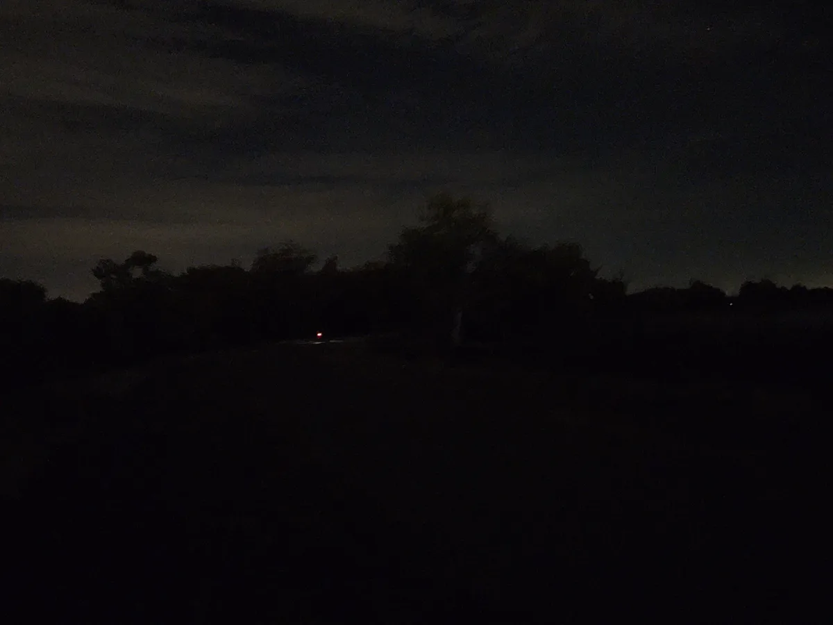 A very dark nighttime photo showing a faint red light in the distance on a road, surrounded by silhouettes of trees against…