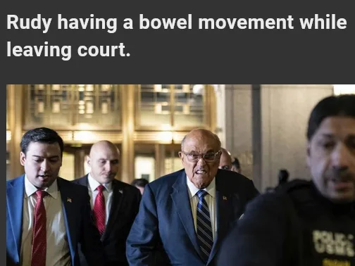 An older man in a suit and tie, flanked by men in suits and a police officer, walks through a courthouse hallway with a…