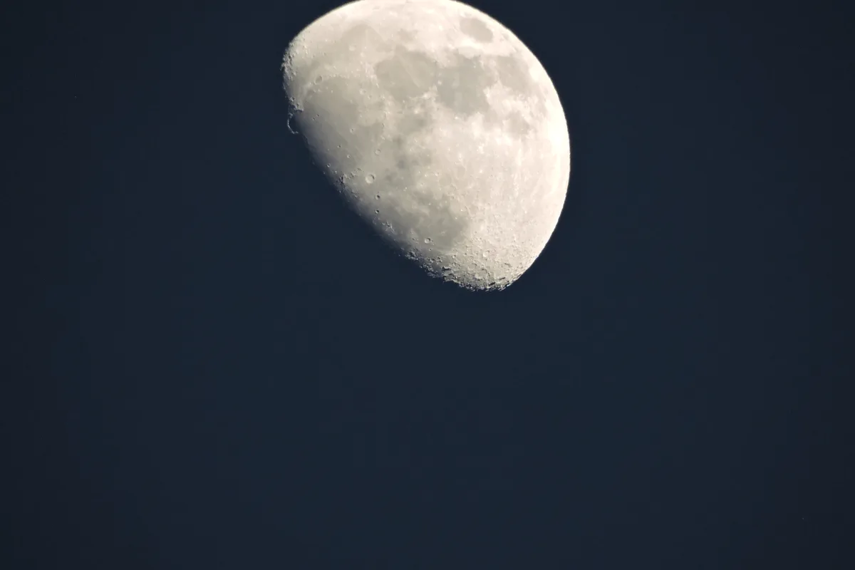 A close-up photograph shows a large, partially illuminated moon against a dark blue sky, with its cratered surface clearly…