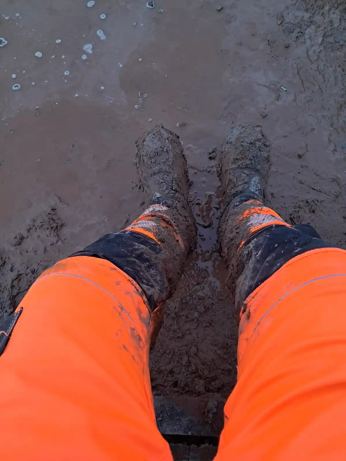 A first-person view shows a person's legs and muddy boots standing in thick, brown mud. They are wearing bright orange…