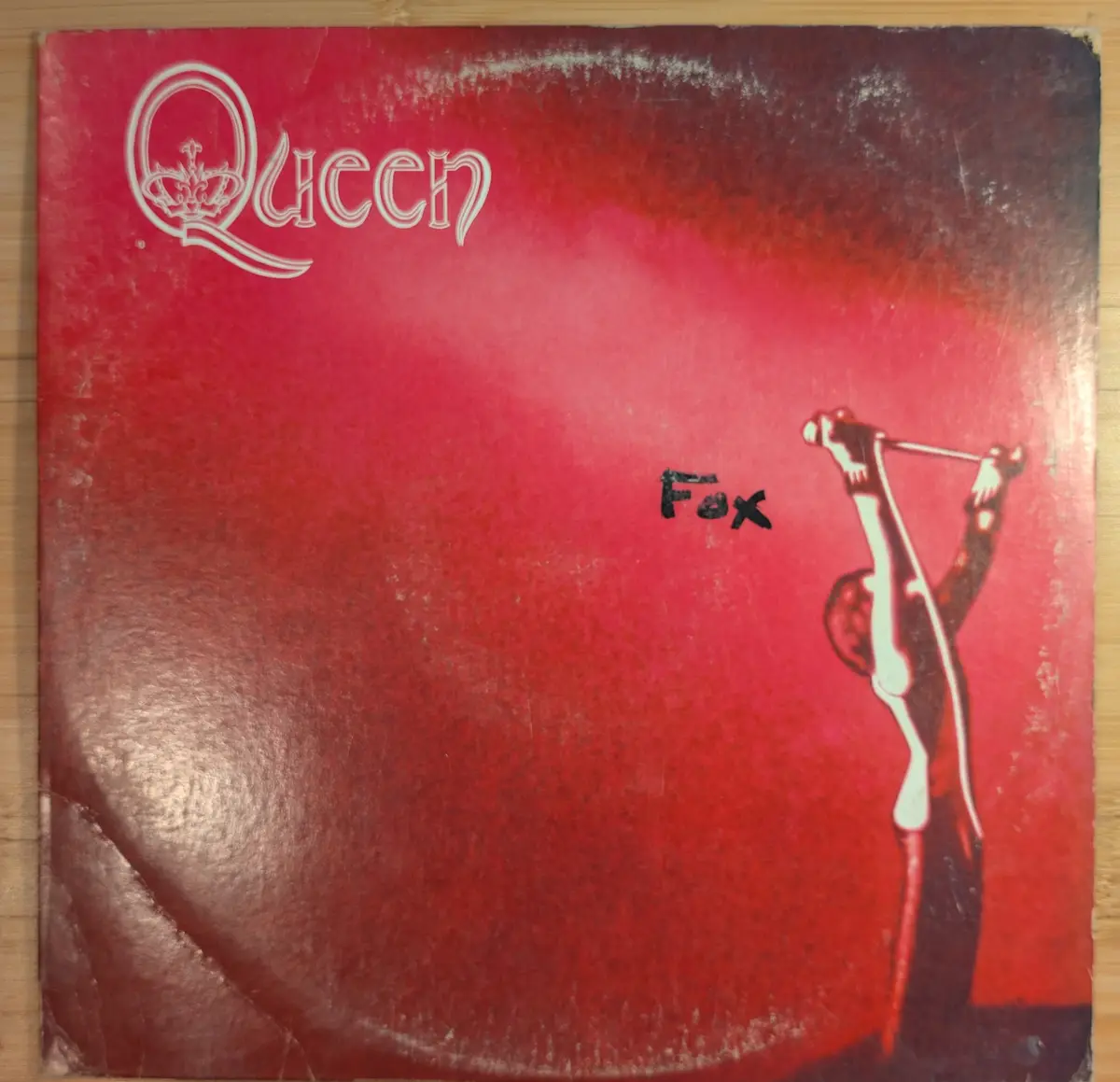 A worn, red Queen album cover featuring the band's logo in white at the top left and a stylized white silhouette of a person…