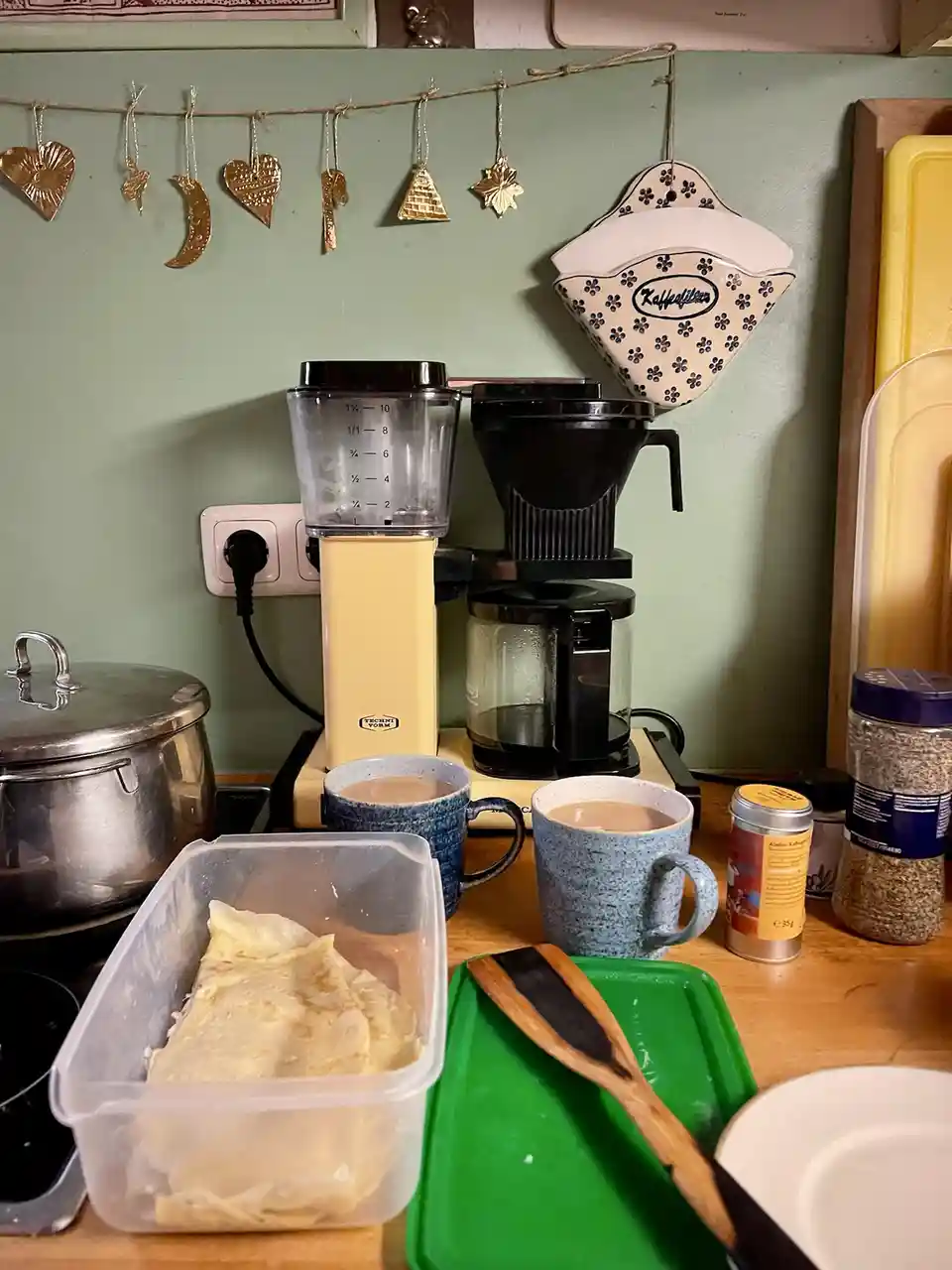 A kitchen counter shows two mugs of coffee, a container of crepes, a coffee maker, and various kitchen items. Above, gold…