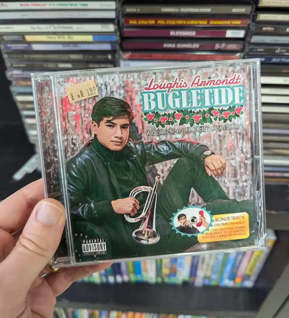 A hand holds a CD case for 'Loughis Anmondt Bugletide: A Collection of Holiday Horn Ditties' in a store, featuring a man in…