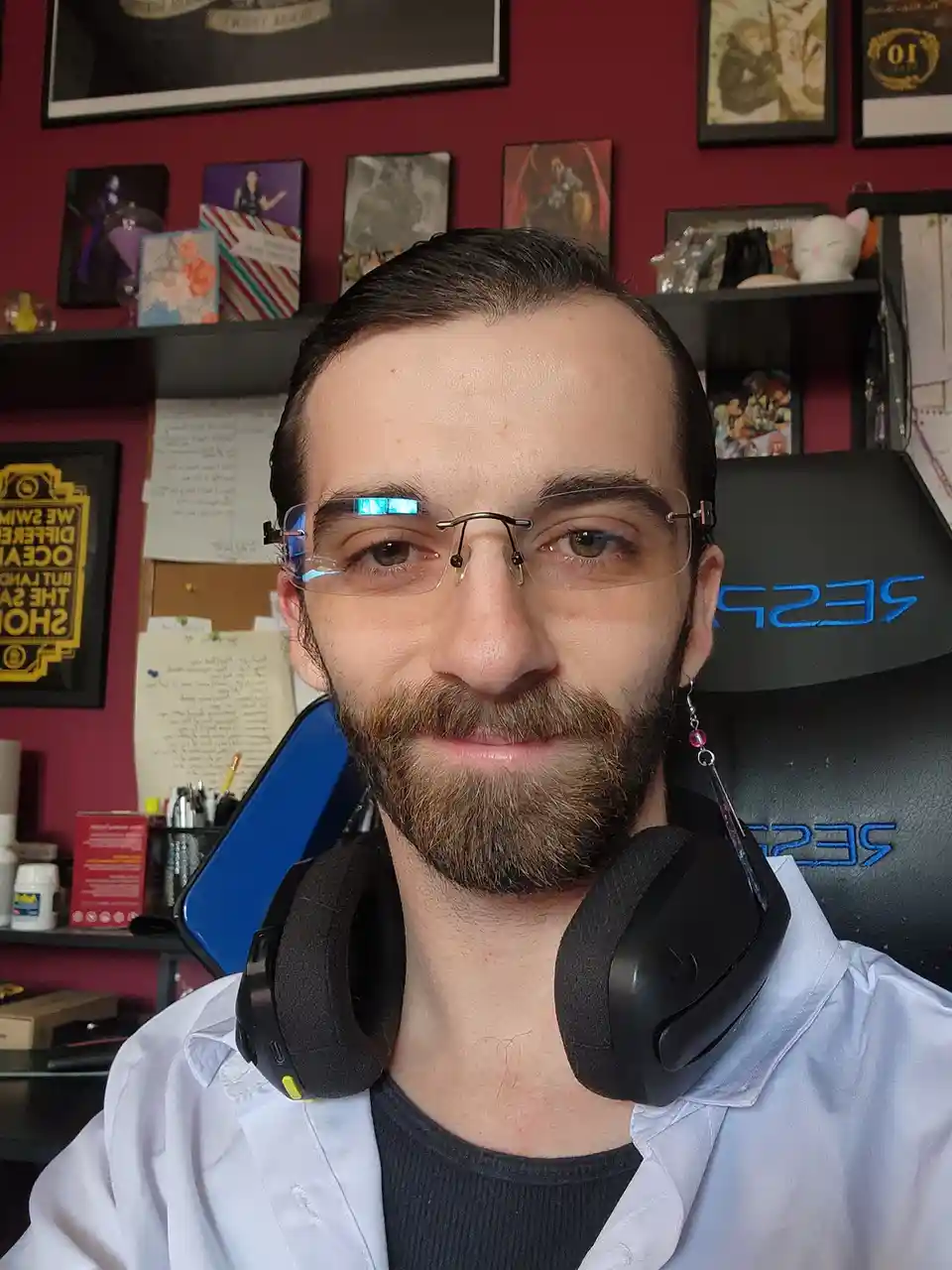 A man with a beard and glasses, wearing a white shirt and black headphones around his neck, sits in a gaming chair. Behind…