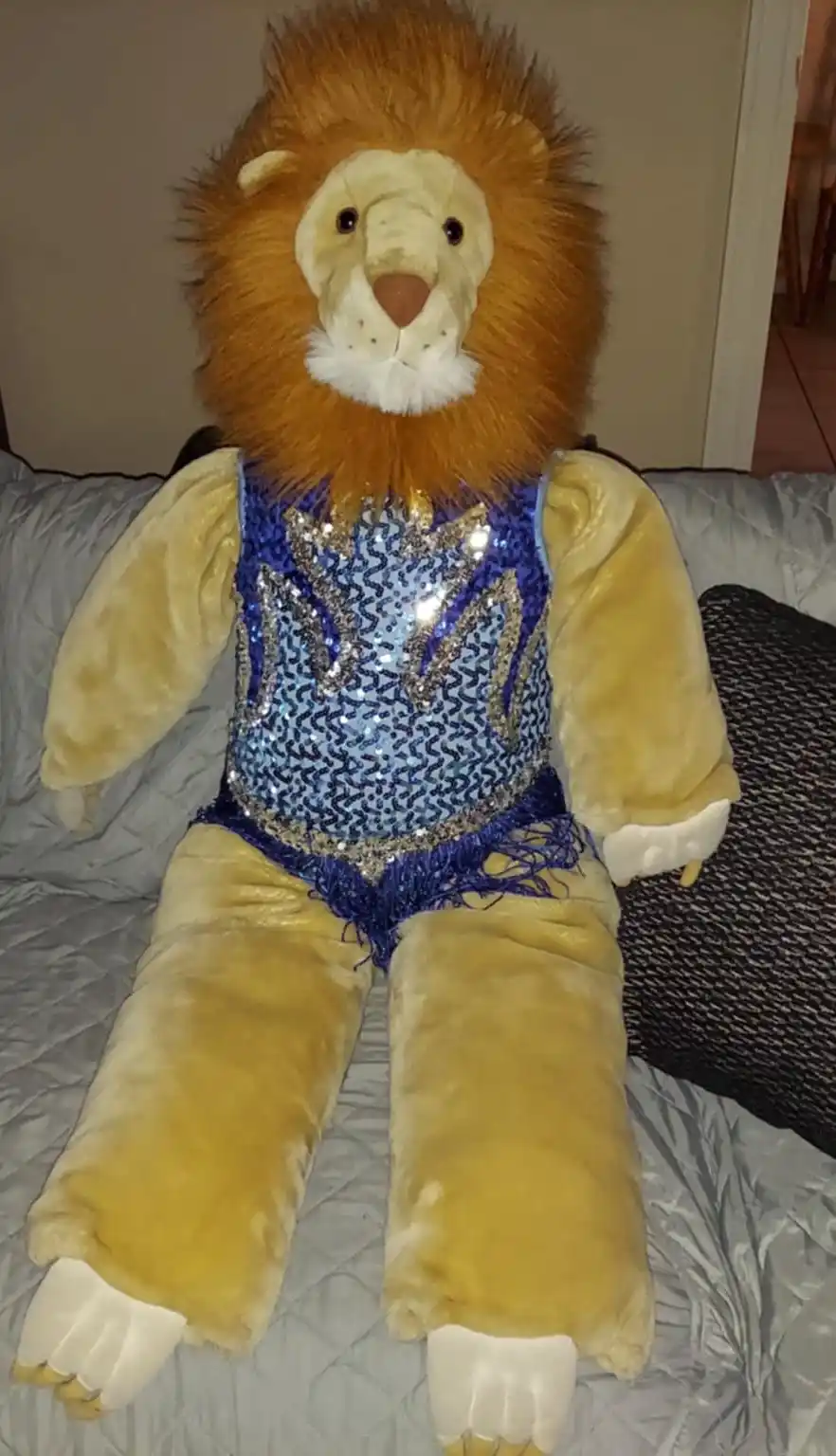 A large, plush lion toy with a shaggy orange mane and tan fur is seated upright on a gray quilted couch. It wears a sparkly…