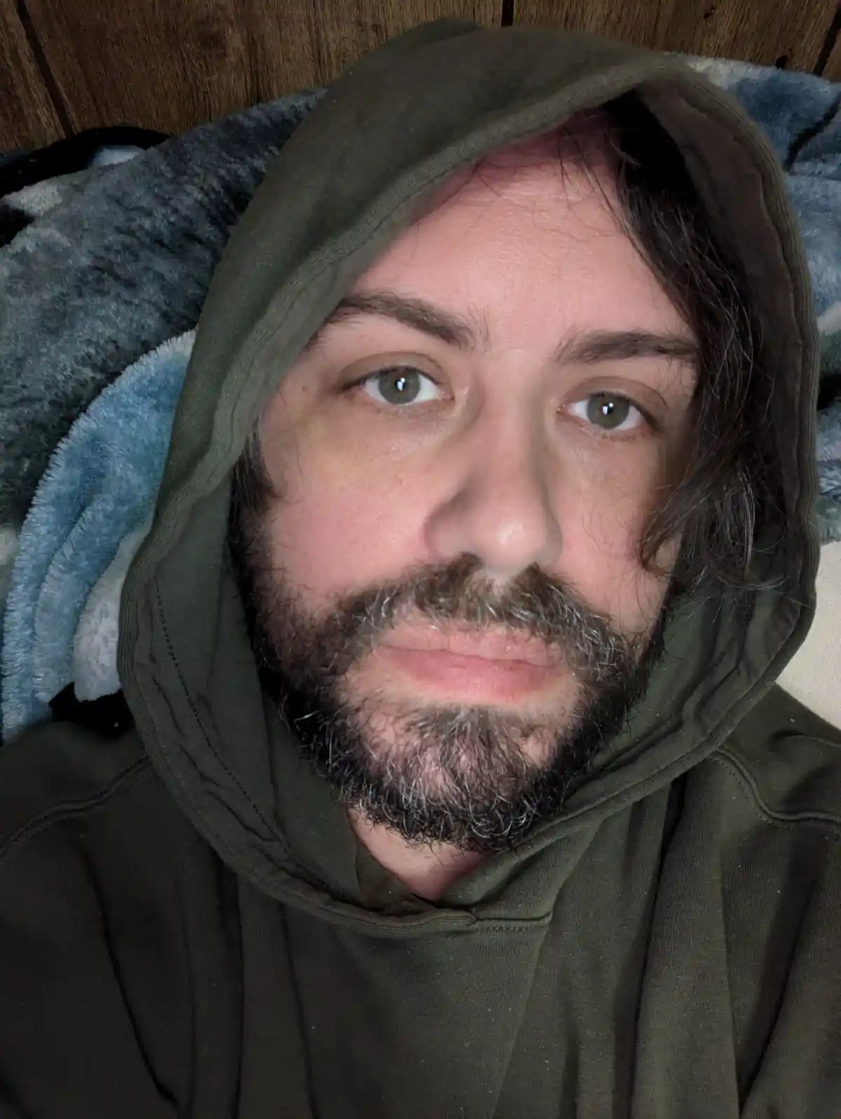A man with a beard and long dark hair wearing an olive green hoodie, looking directly at the camera with a neutral…