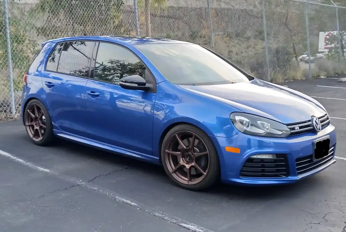 A blue Volkswagen Golf R with bronze-colored wheels is parked in an asphalt lot next to a chain-link fence. The car's glossy…