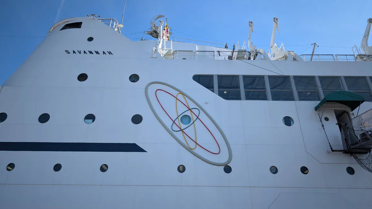 A low-angle photo shows the white hull of a ship named 'SAVANNAH' under a clear blue sky. The ship features round portholes…