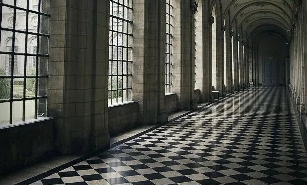 A long, empty, grand hallway with tall stone columns and large windows, featuring a glossy black-and-white checkered floor…
