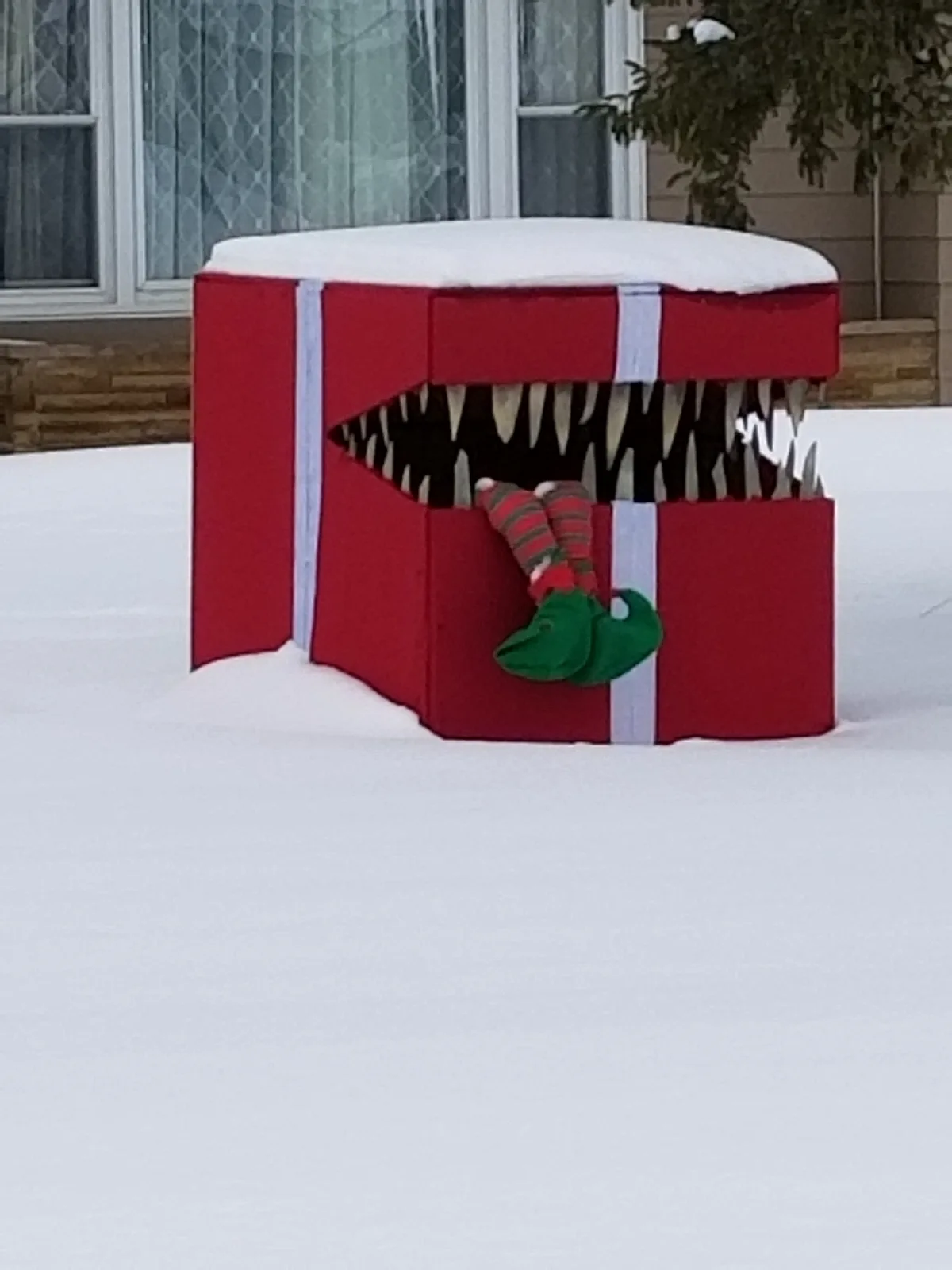 A large red gift box with white ribbon sits in a snowy yard, its top open to reveal a monstrous mouth full of sharp teeth. A…