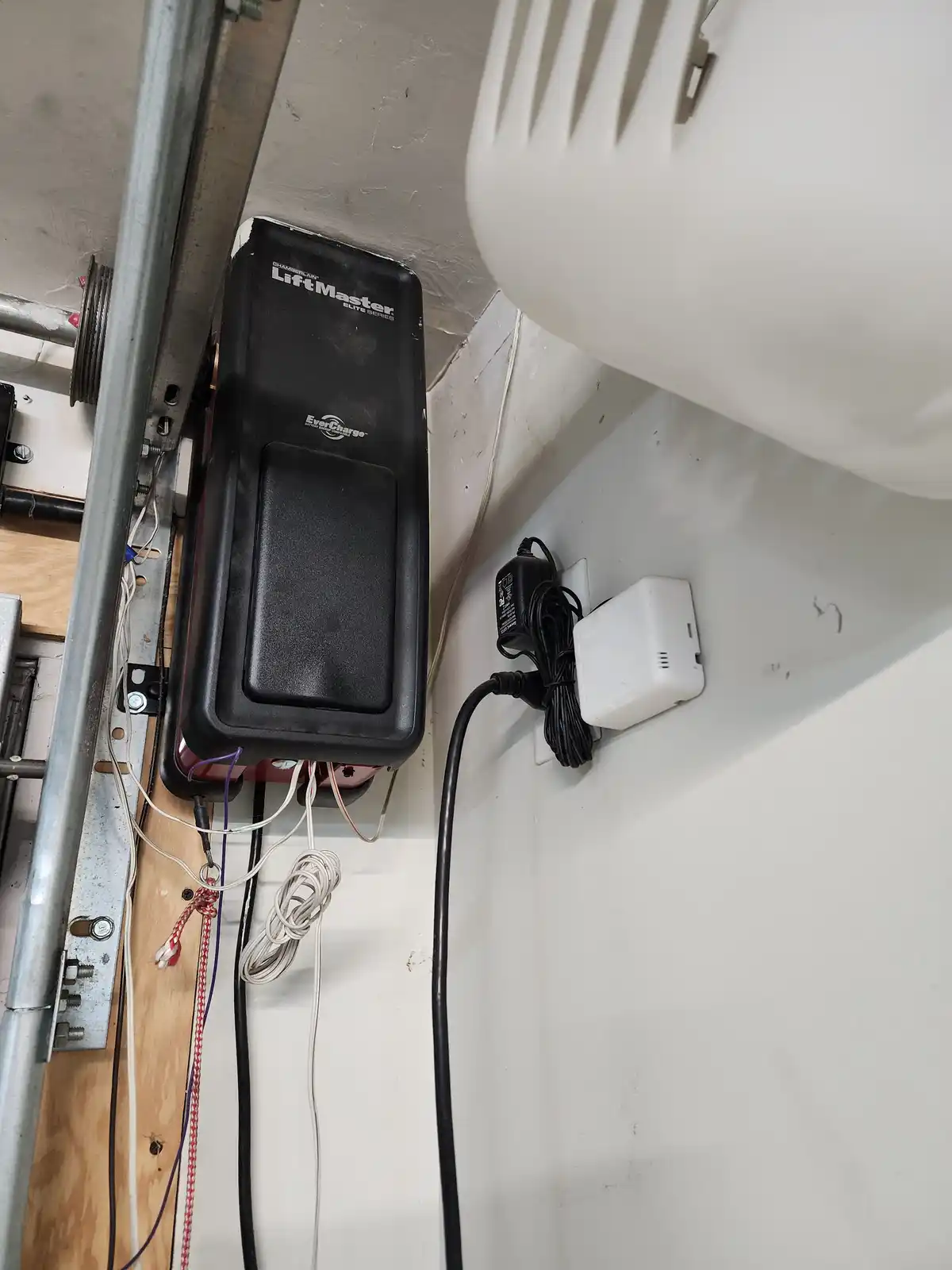 A black Chamberlain LiftMaster Elite Series garage door opener is mounted on a wall next to a white plastic vent, with…