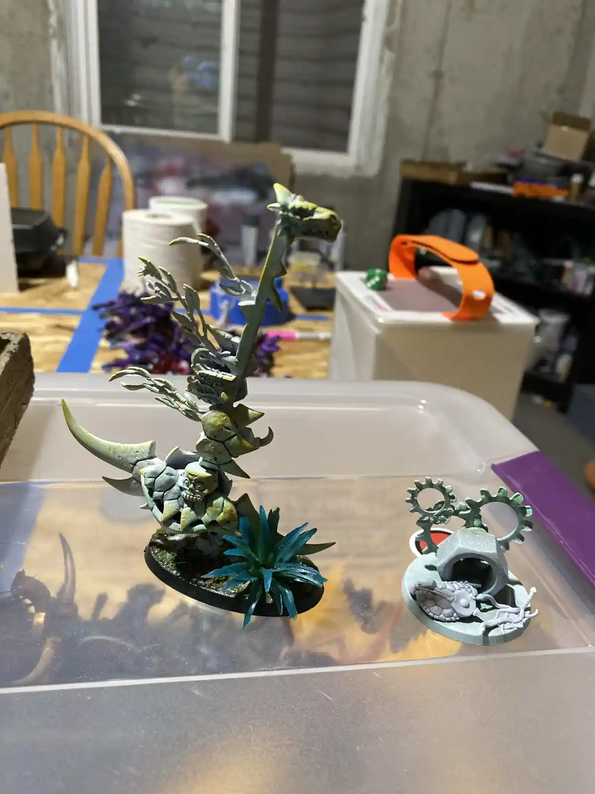 A photo shows two painted miniature figurines on a clear plastic tray. The larger one is a greenish, ornate creature with…