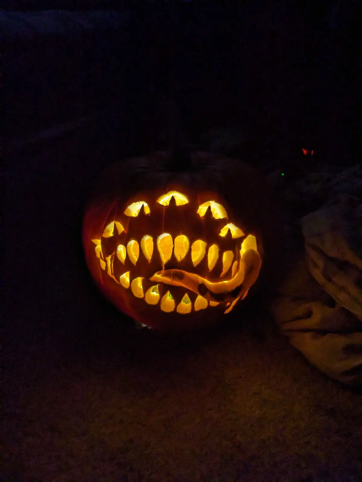 A carved Halloween pumpkin with a wide, toothy grin and pointed ears, illuminated from within in a dark setting. The…