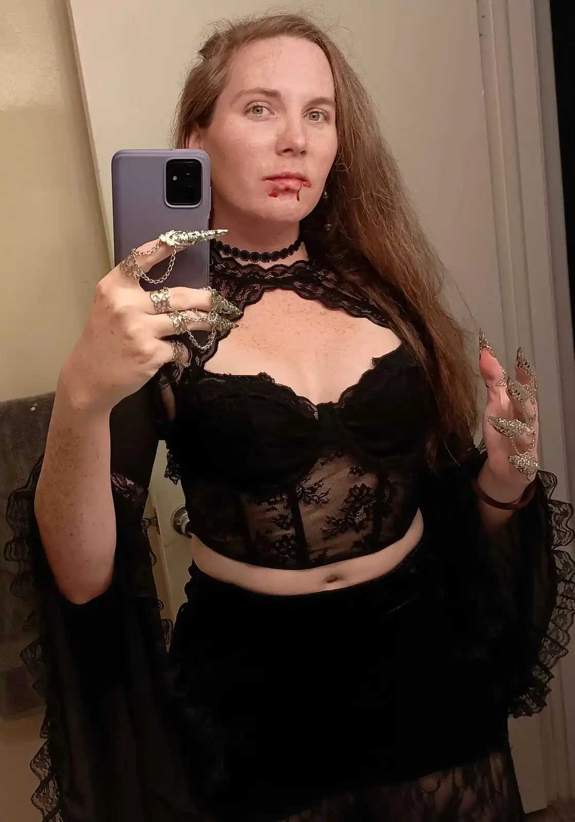 A woman with long brown hair takes a mirror selfie holding a purple phone, wearing a black lace and velvet gothic outfit…
