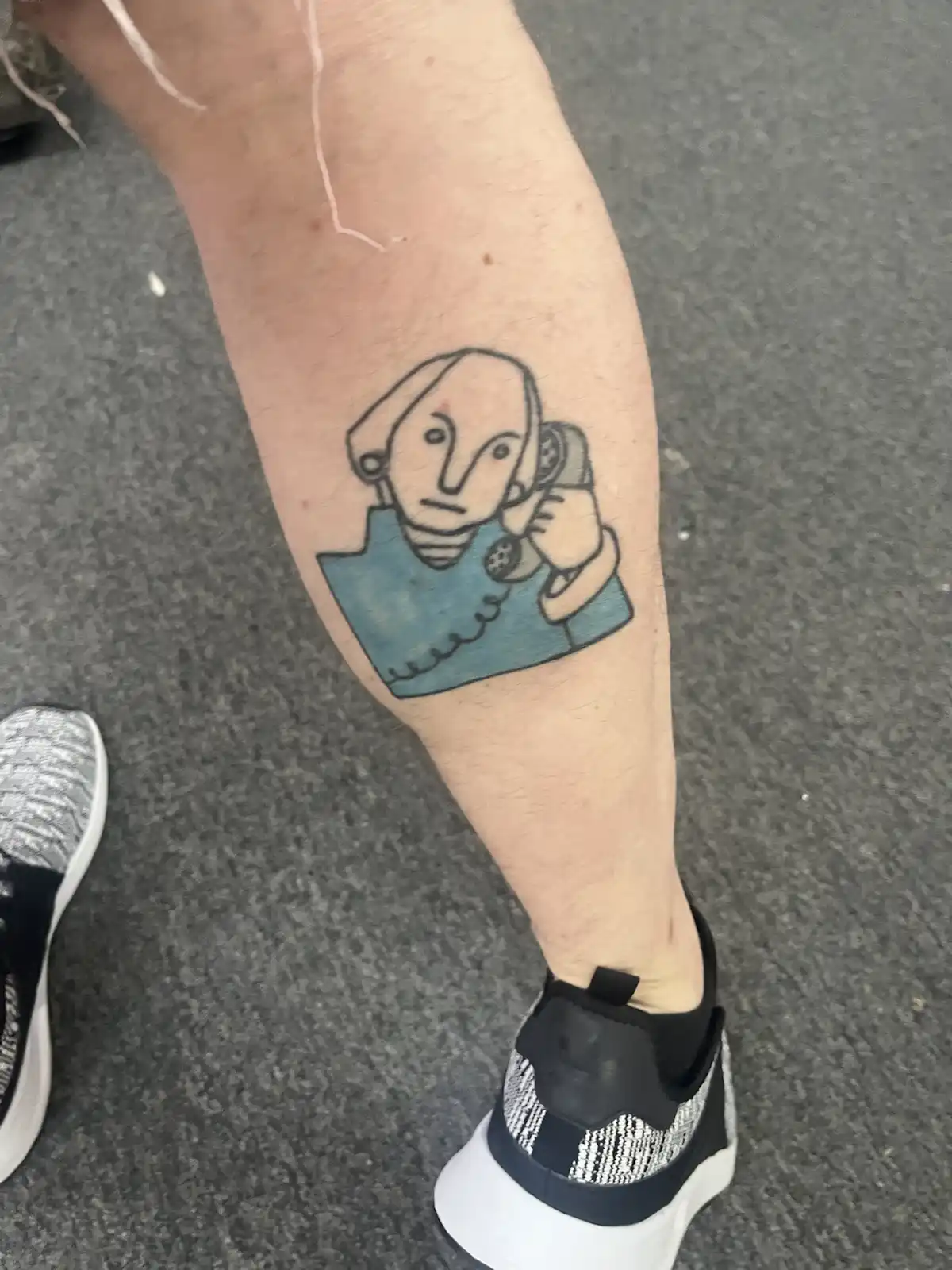 A close-up photo shows a tattoo on a person's lower leg, depicting a simple cartoon-style figure with a bald head, wearing a…