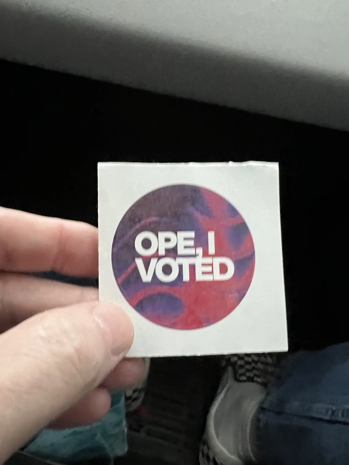 A hand holds up a square sticker with a circular design featuring red and purple swirls. The sticker has bold white text…