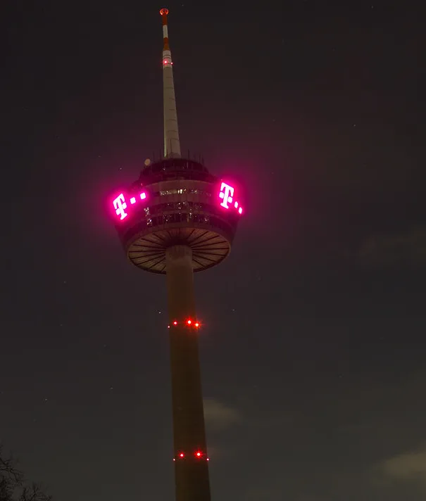 A tall tower at night with red lights spelling out 't-mobile' on its circular observation deck. The tower has a slender…