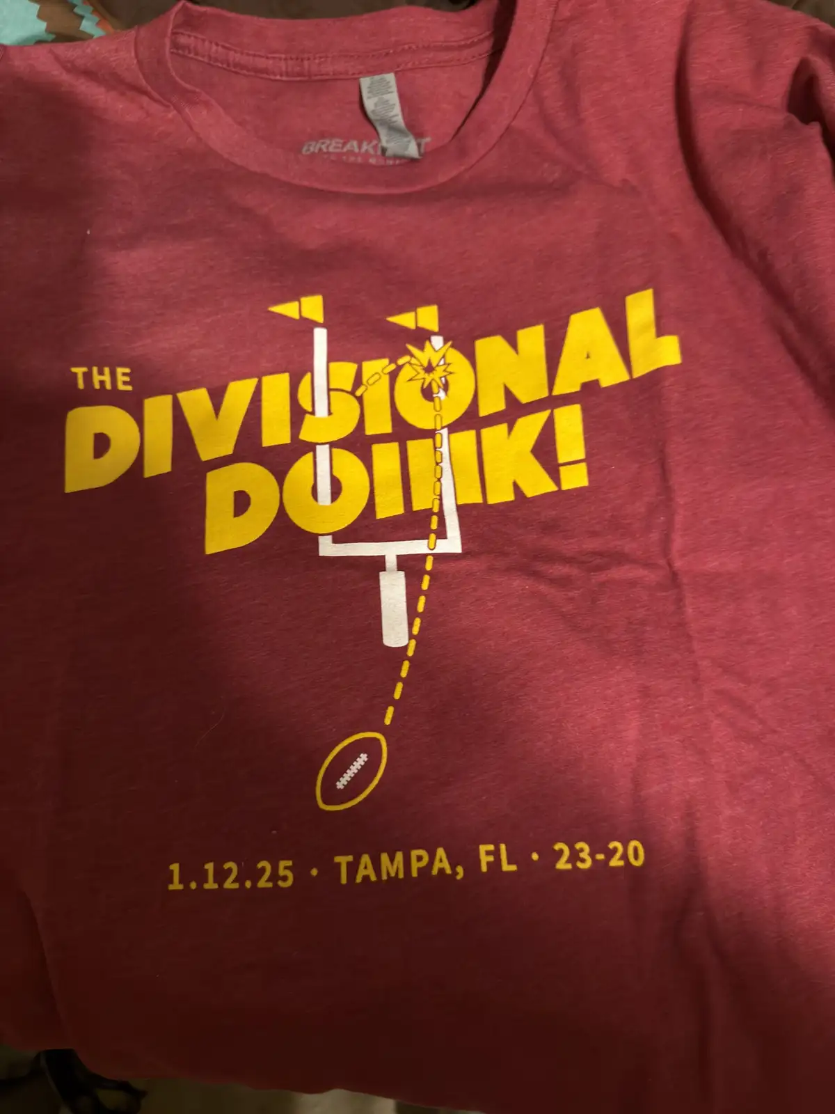 A close-up of a maroon t-shirt with yellow text and graphics. The text reads 'THE DIVISIONAL DOINK!' with a graphic of a…