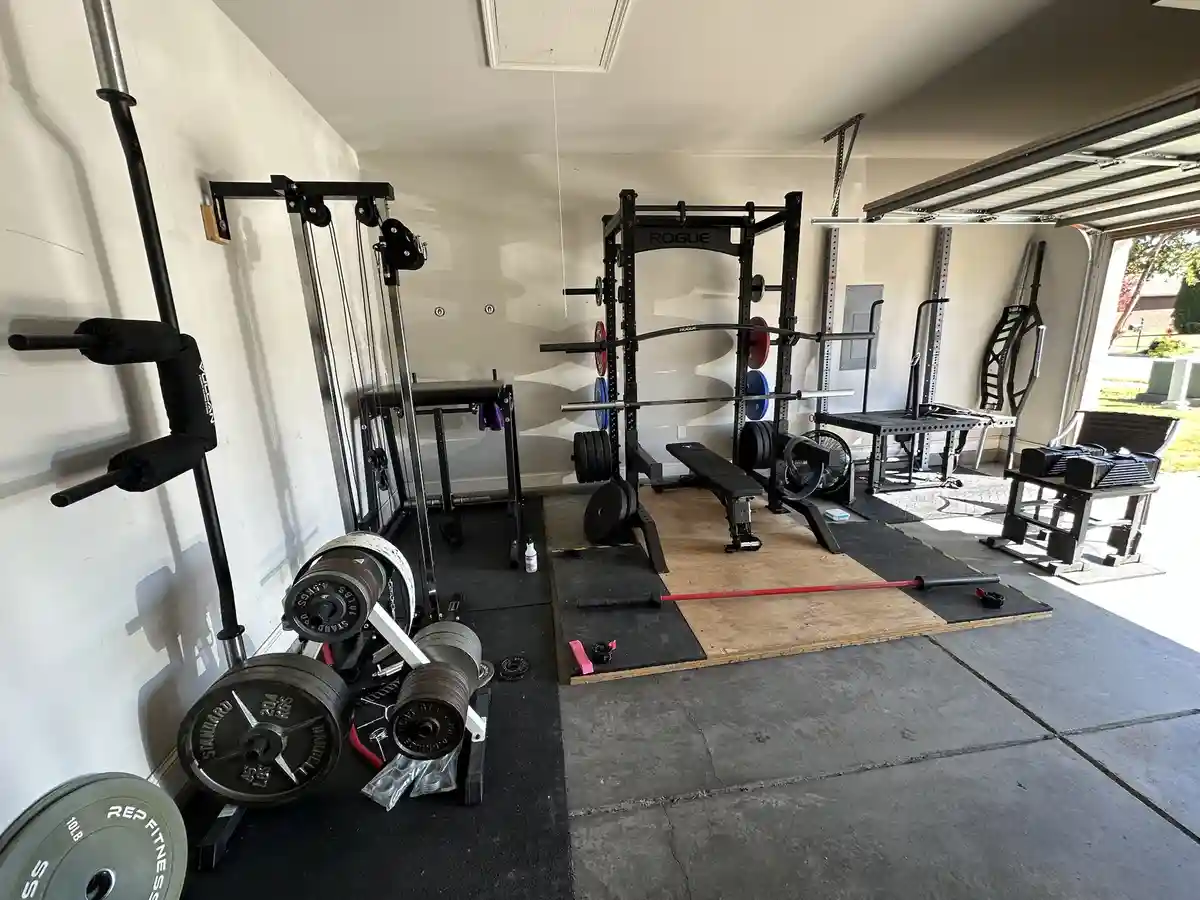 A well-equipped home gym set up in a garage, featuring a Rogue squat rack with weights, a cable machine, barbells, weight…