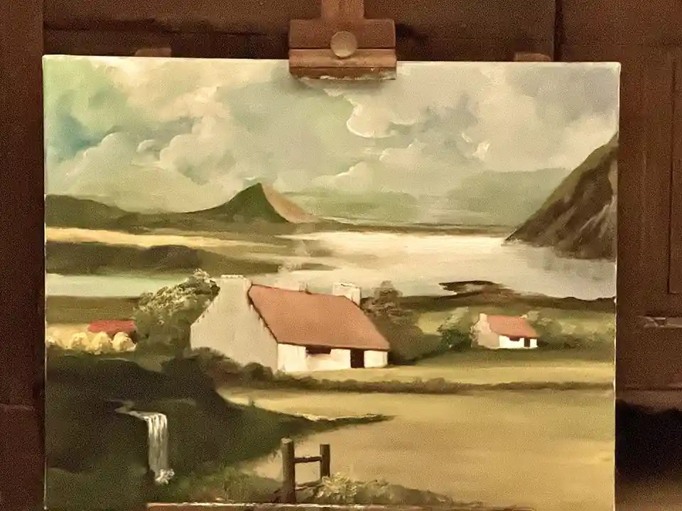 A painting of a rural landscape with white cottages, rolling hills, and a river is displayed on an easel. The sky in the…