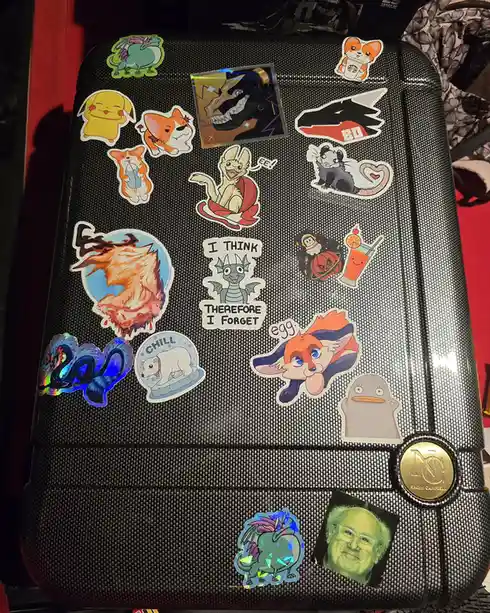 A black textured suitcase is covered in numerous colorful stickers featuring cartoon animals, anime characters, and pop…