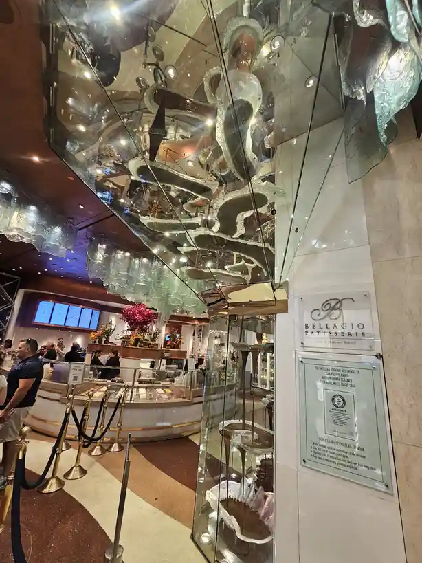 A large, multi-tiered chocolate fountain is enclosed in glass and suspended from the ceiling inside Bellagio Patisserie…