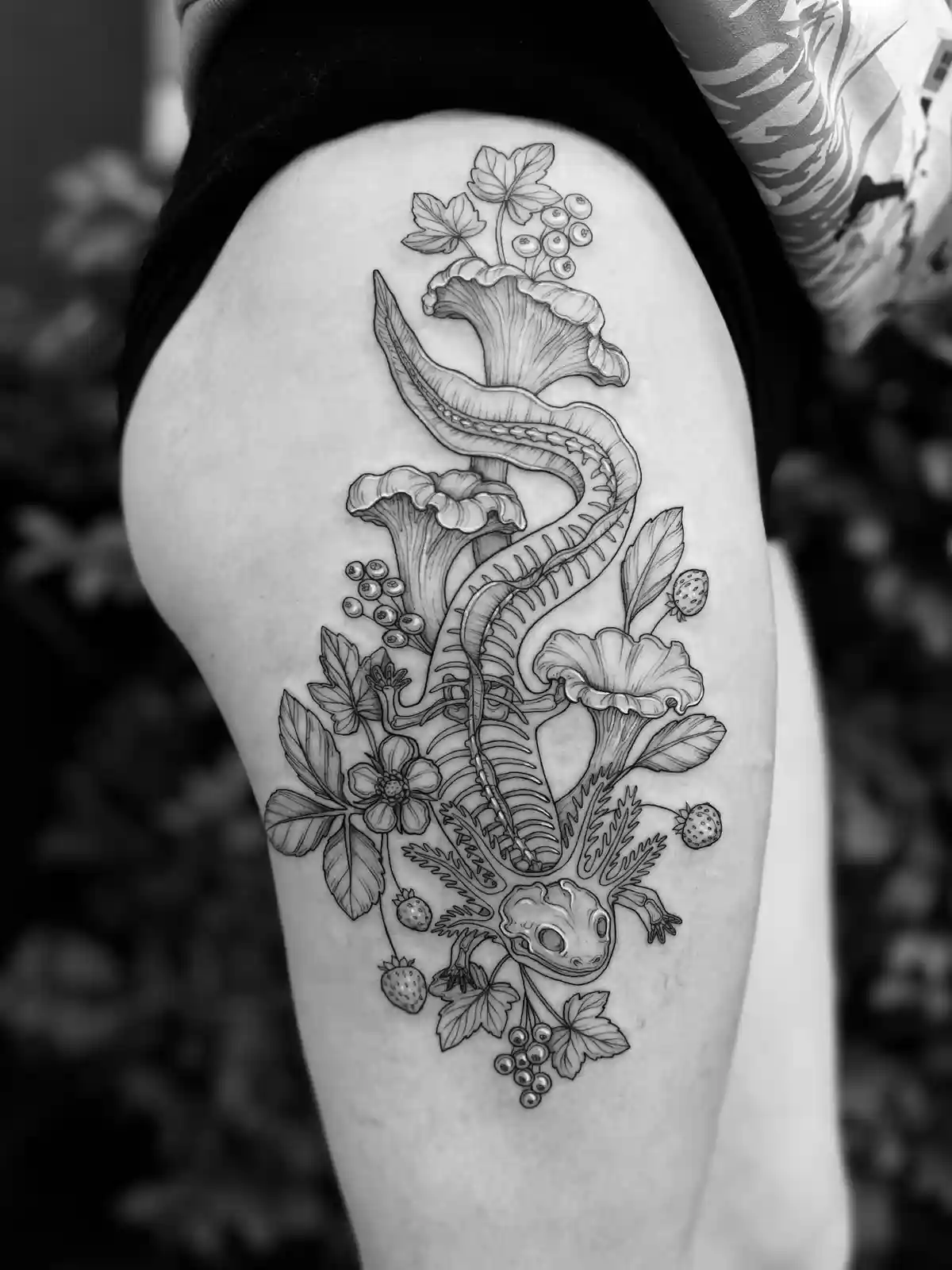 A black-and-white photo shows a detailed tattoo on a person's thigh, featuring a skeletal newt or salamander intertwined…
