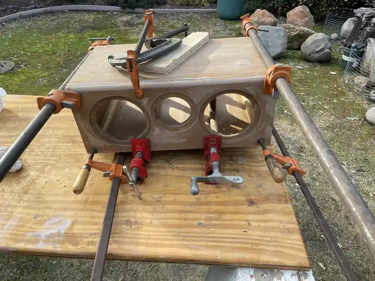 A wooden box with three circular cutouts is clamped heavily with multiple orange and red clamps on a workbench outdoors. The…